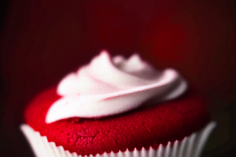 A close-up of a gluten-free red velvet cupcake under bright, clear studio lighting, filling the frame and in razor-sharp focus on the frosting and crumb texture