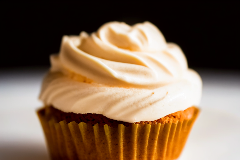 A gluten-free pumpkin spice cupcake on a white plate under bright, directional studio lighting with razor-sharp focus on the cream cheese frosting, filling the frame