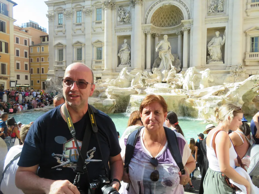 A photo of a man and a woman standing by the Trevi Fountain in Rome. The water ripples and moves behind them