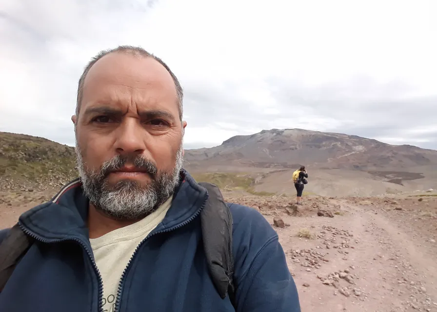 a man standing on a dirt road with mountains in the background. sky, cloud, mountain, beard, travel, mountainous landforms, jacket, landscape, wrinkle, hill