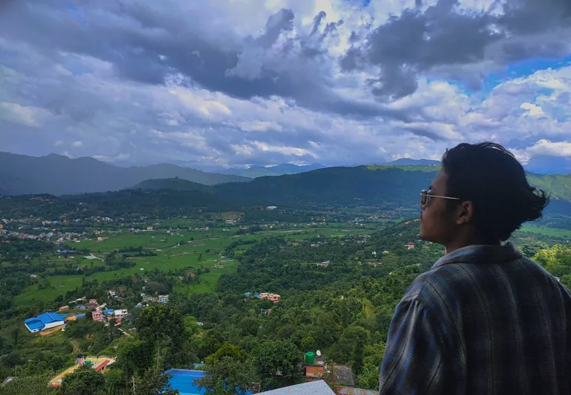 A man standing on top of a lush green hillside, with a beautiful blue sky behind him. moving a clouds