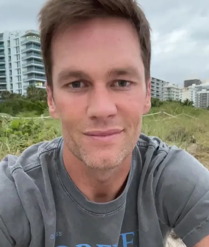 a man in a gray shirt sitting on a beach. forehead, nose, hair, cheek, hairstyle, daytime, eyebrow, plant, jaw, sky