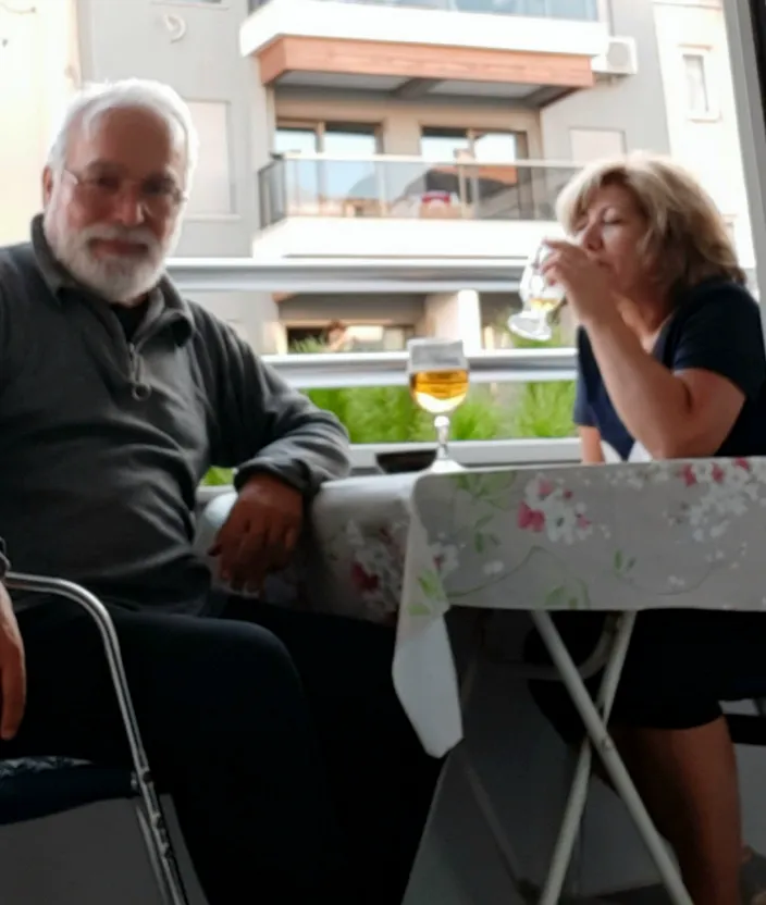 a man sitting next to a woman at a table. table, window, tableware, t-shirt, people, building, outdoor table, leisure, beard, fun