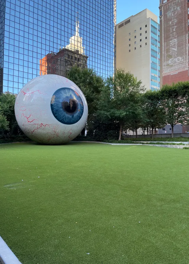 a large eyeball sitting on top of a lush green field. building, sky, plant, skyscraper, architecture, tree, ball, grass, tower block, art