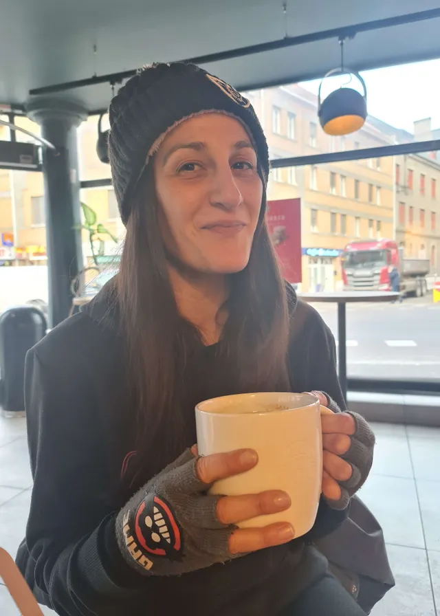 a woman sitting at a table holding a cup of coffee. smile, cup, eyewear, finger, cool, vehicle, window, drink, automotive design, serveware