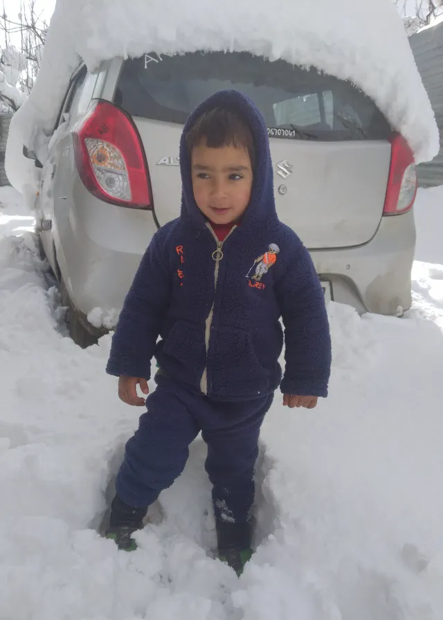 a little boy standing in the snow next to a car. car, snow, land vehicle, wheel, tire, vehicle, white, automotive tire, automotive lighting, motor vehicle