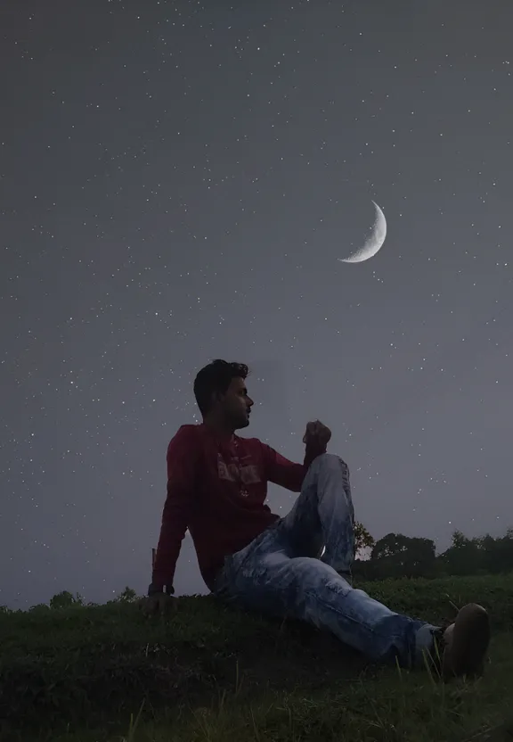 a man sitting in the grass with a half moon in the background