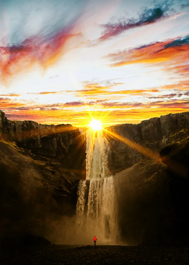 a person standing in front of a waterfall at sunset. cloud, water, sky, atmosphere, water resources, ecoregion, mountain, natural landscape, orange, sunlight