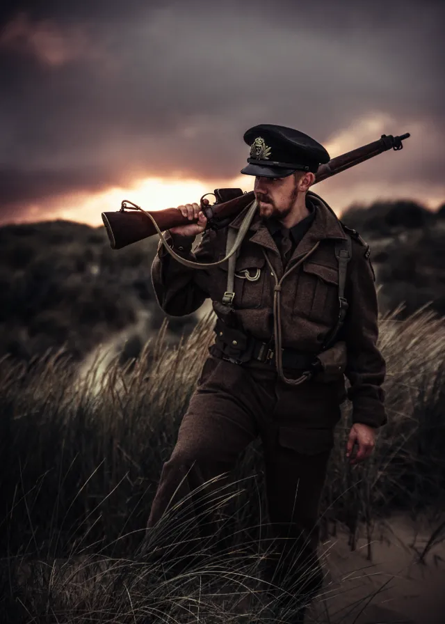 a man in a uniform with a gun in his hand. outerwear, hat, sky, cloud, flash photography, plant, people in nature, grass, grassland, landscape