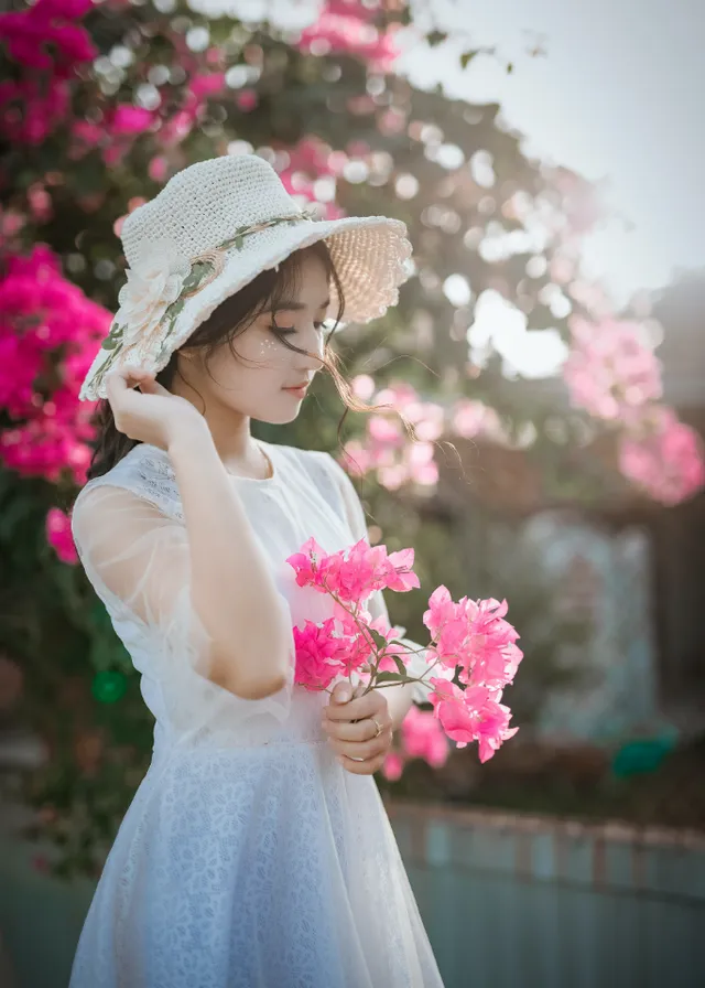 a woman in a white dress holding pink flowers. clothing, face, flower, head, lip, plant, hairstyle, shoulder, white, petal