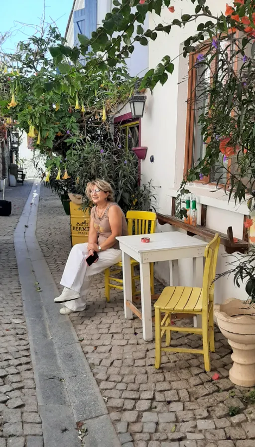 a woman sitting on a yellow chair next to a table. building, table, flowerpot, houseplant, house, outdoor furniture, plant, outdoor table, leisure, tree